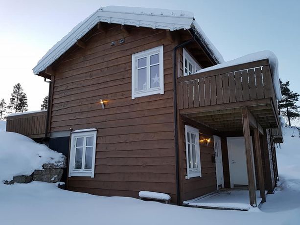 Hytte dekt i snø i et vinterlandskap med hvite vinduer og hvite dekortert takskjegg samt julestjerner i vinduene