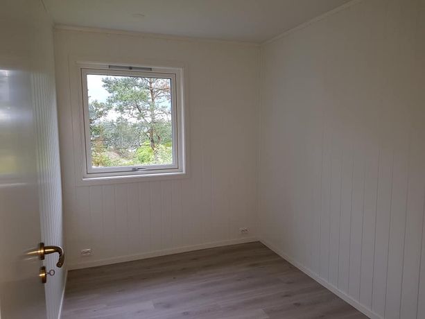 Et lite rom med hvitmalte trevegger, parkett gulv med tre mønster, et vindu med hvite vinduskarmer med utsikt til trær