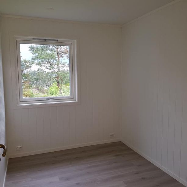 Et lite rom med hvitmalte trevegger, parkett gulv med tre mønster, et vindu med hvite vinduskarmer med utsikt til trær