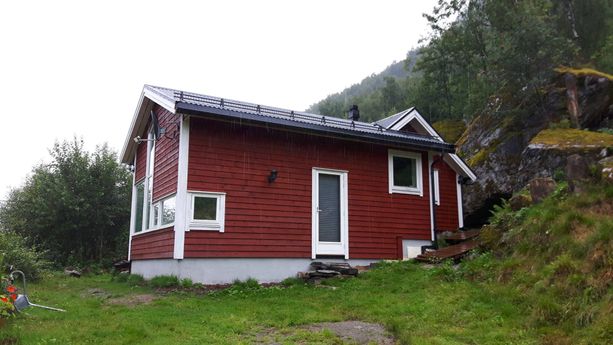 En rød hytte med en liten steintrapp opp til en dør og mange mindre vindu med hvite vinduskarmer som står ved foten av et fjell