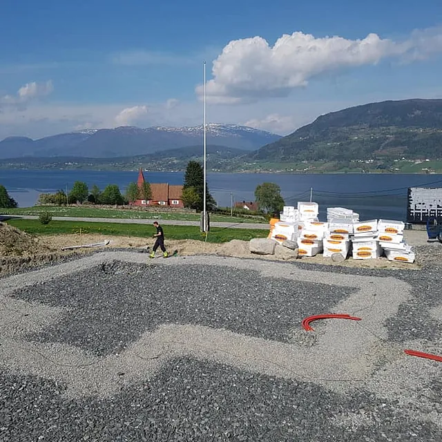 Oppstart betongarbeid med en ansatt i bakgrunnen med en haug med oppgravd jord på ene siden og masse utstyr til det nye huset på høyre siden ved siden av to firmabiler