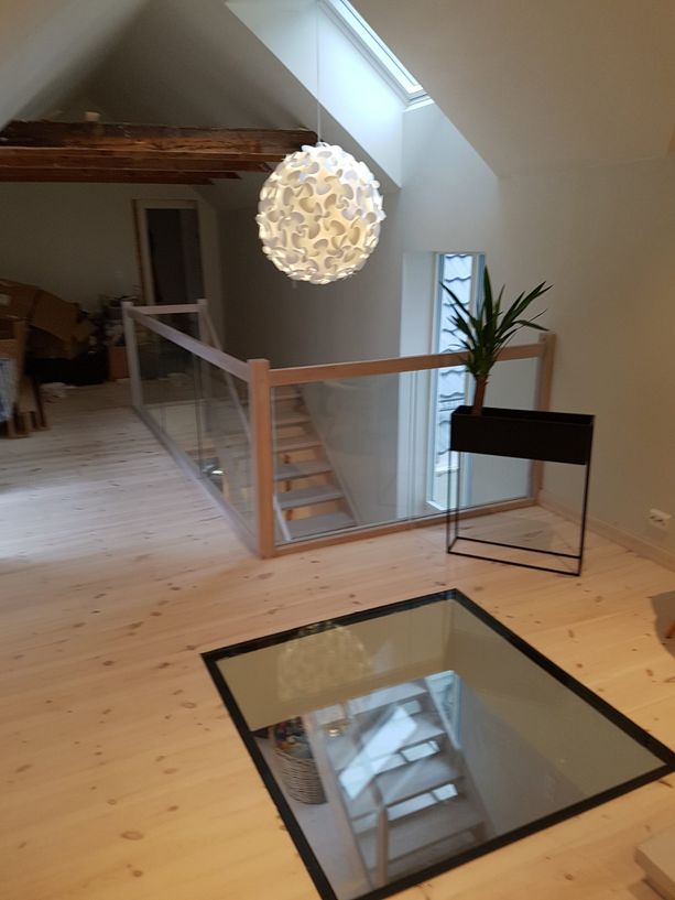 Et takvindu i 2 etasje som har en blomsterpotte, taklampe synlige bjelker, glass gelender rundt trappen og et lyst og moderne interiør med vindu som gir innsyn i etasjen under hvor man ser begynnelsen på trappen