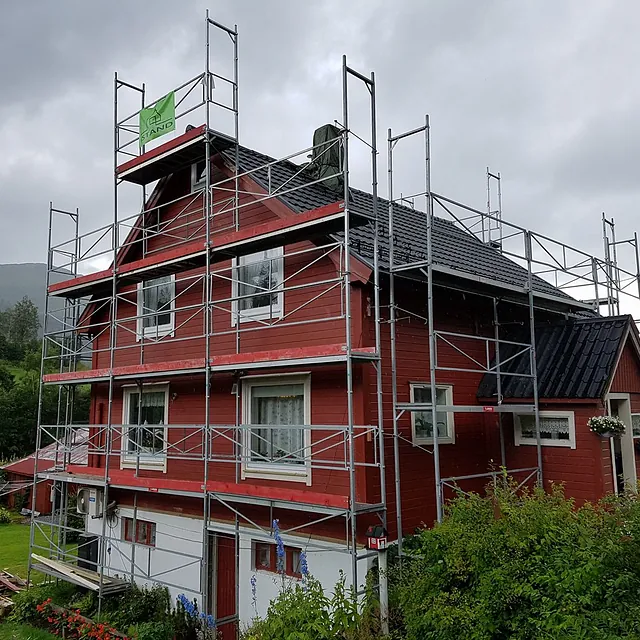 Stilas satt opp et rødt hus med iStand AS logo på for å bytte kledning og tak med en grå sky og fjell ibakgrunnen