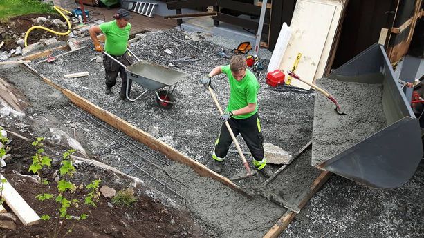 To ansatte som støper fundament for et tilbygg med trillebårer, verneutstyr, spader og en traktor skuff fylt med støpe fundament