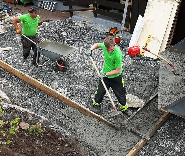 To ansatte som støper fundament for et tilbygg med trillebårer, verneutstyr, spader og en traktor skuff fylt med støpe fundament