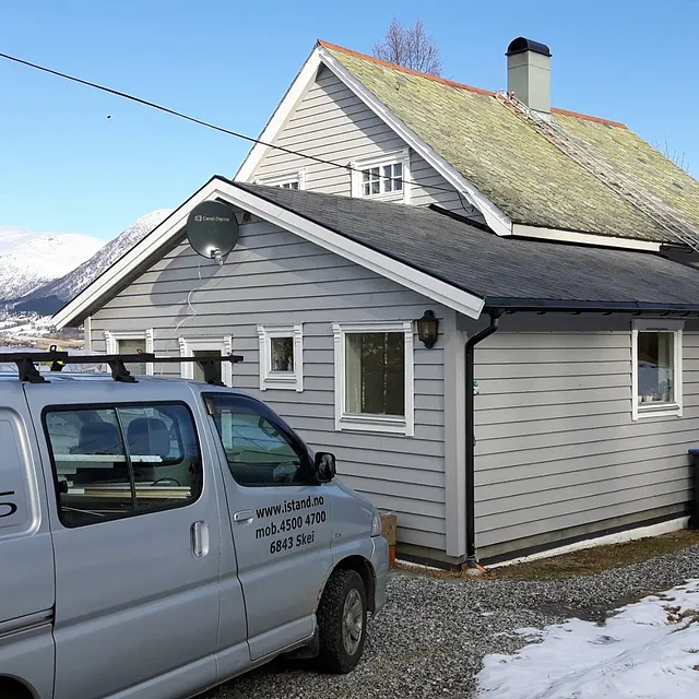 En firmabil som står parkert utenfor et grått hus med et tilbygg og det originale bygget med grønt tak