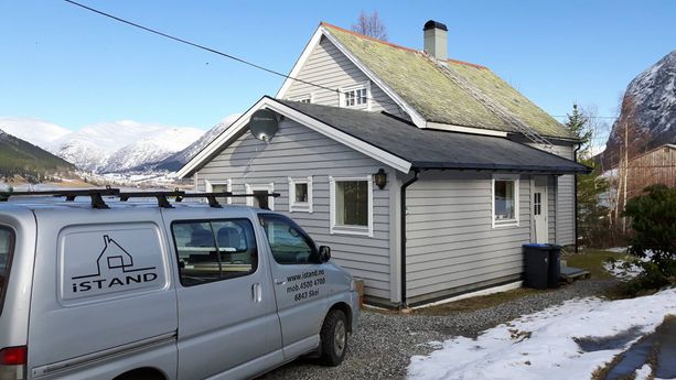 En firmabil som står parkert utenfor et grått hus med et tilbygg og det originale bygget med grønt tak