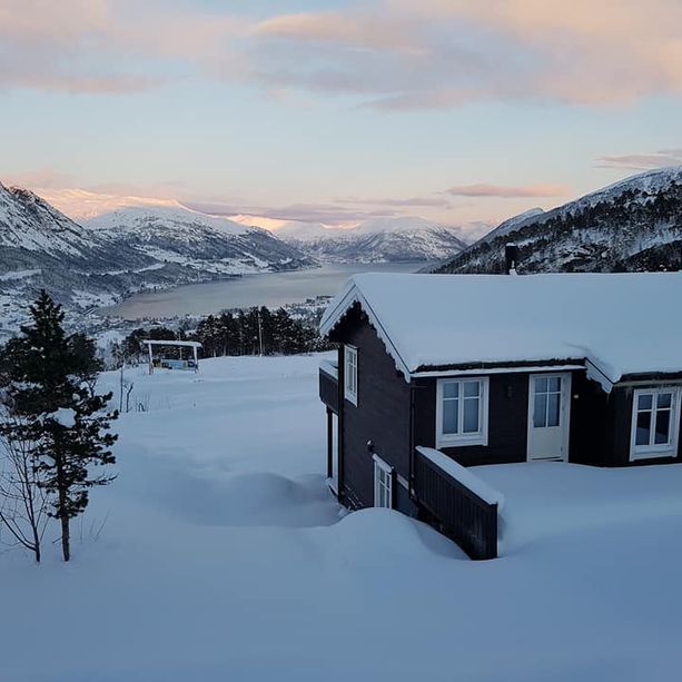 En mørk hytte i et vinterlandskap dekket av sne med hvite vinduer og dører og utsikt over en fjord og fjell