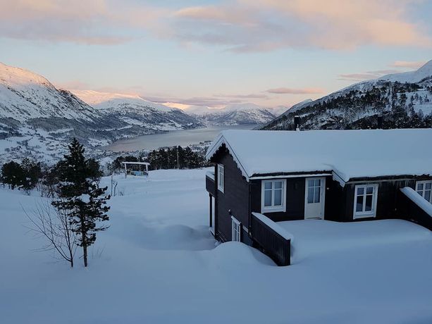 En mørk hytte i et vinterlandskap dekket av sne med hvite vinduer og dører og utsikt over en fjord og fjell
