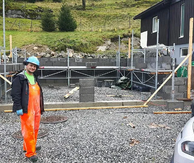 En ansatt med hjelm, øreklokker og verneklær på seg som står forran grunnmuren til en garasje hvor noe av veggen har blitt satt opp med stilas på innsiden av garasjen gvor man også ser firmabiler i høyre hjørne