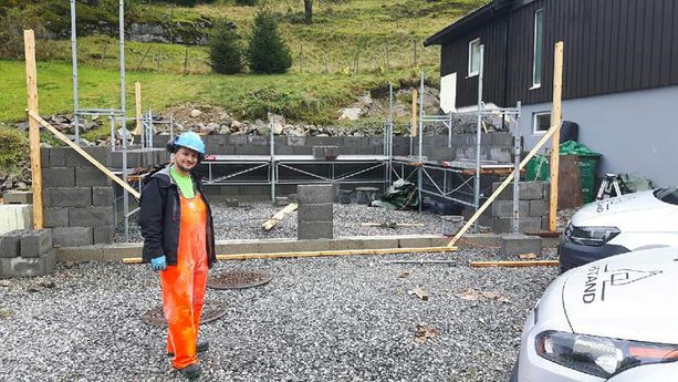 En ansatt med hjelm, øreklokker og verneklær på seg som står forran grunnmuren til en garasje hvor noe av veggen har blitt satt opp med stilas på innsiden av garasjen gvor man også ser firmabiler i høyre hjørne