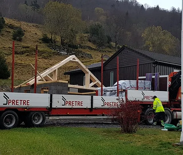 Prosessen med å bygge en garasje til et hus med en lastebil med en kran i forgrunnen hvor en arbeider går og sjekker lastebilen 