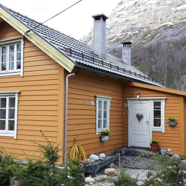 En oransje hytte med hvite vindu, skifertak, hvit inngangsdør, to piper som stikker opp fra taket og et stort fjell i bakgrunnen 