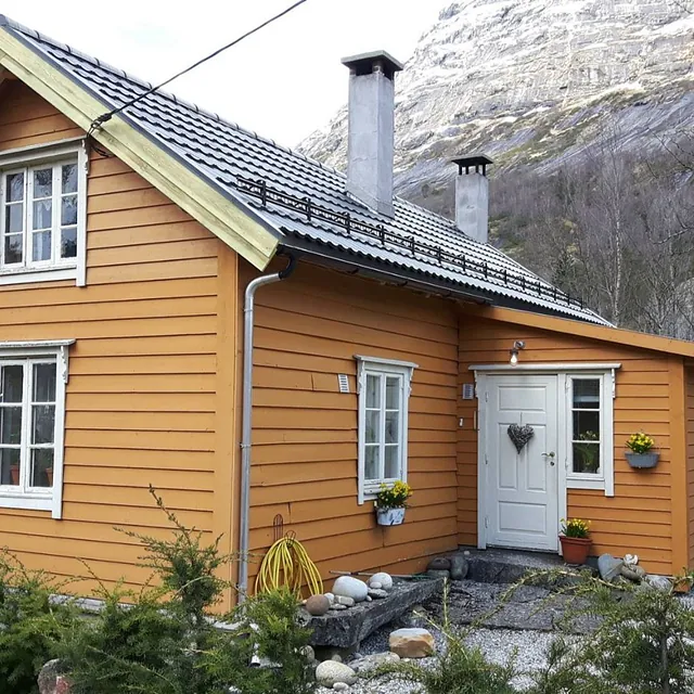En oransje hytte med hvite vindu, skifertak, hvit inngangsdør, to piper som stikker opp fra taket og et stort fjell i bakgrunnen 