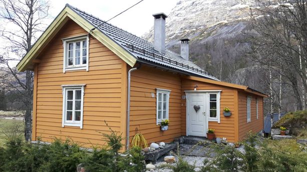 En oransje hytte med hvite vindu, skifertak, hvit inngangsdør, to piper som stikker opp fra taket og et stort fjell i bakgrunnen 