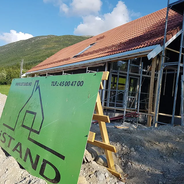 Firmalogo som står satt opp på eiendommen til et helt nytt hus som holder på å bli bygt opp fra grunnen med vegger, vindu og tak ferdig snekret og stilas installert rundt utsiden 