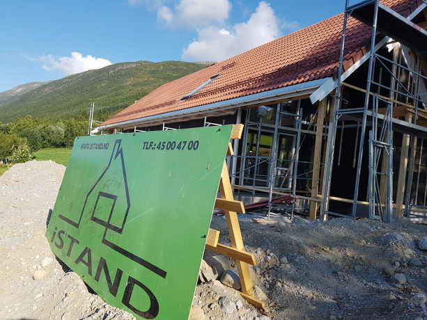 Firmalogo som står satt opp på eiendommen til et helt nytt hus som holder på å bli bygt opp fra grunnen med vegger, vindu og tak ferdig snekret og stilas installert rundt utsiden 