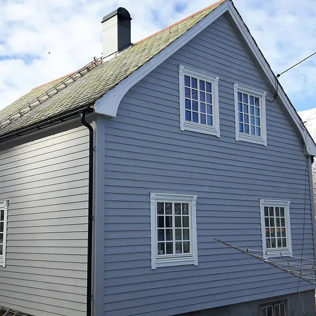 Grått hus med hvite vindu med detaljer på vinduskarmene og tak kant med en pipe som kommer opp fra taket