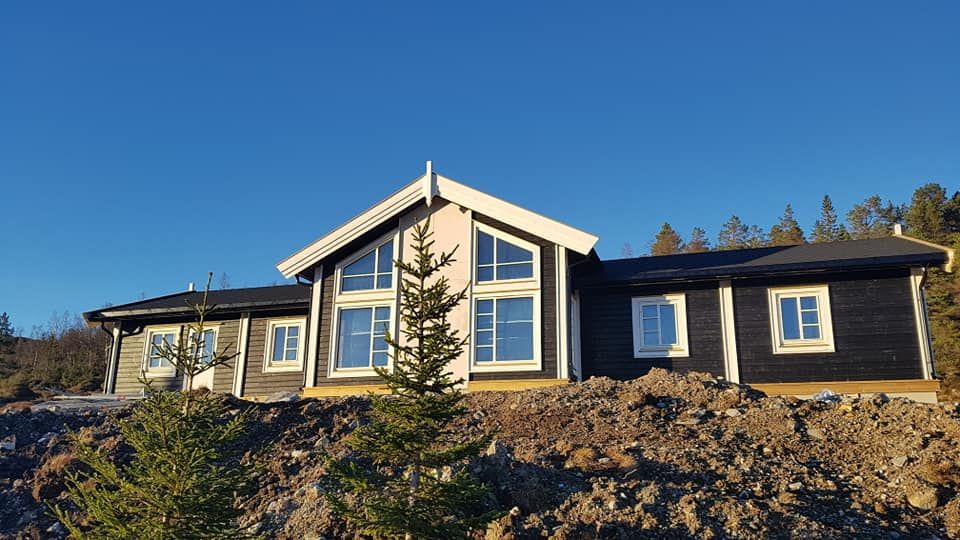 En svart hytte med hvite detaljer på vinduskarmer, dører og takskjegg under en blå hummel med trær i bakgrunnen