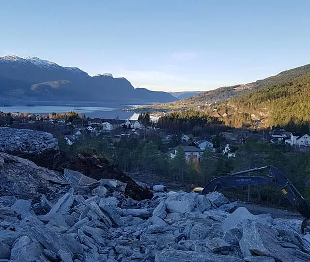 Utsikt fra toppen av en grøft hvor en gravemaskin legger ned stein hvor man ser ut over en bygd med fjord og fjell 
