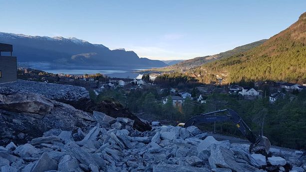 Utsikt fra toppen av en grøft hvor en gravemaskin legger ned stein hvor man ser ut over en bygd med fjord og fjell 