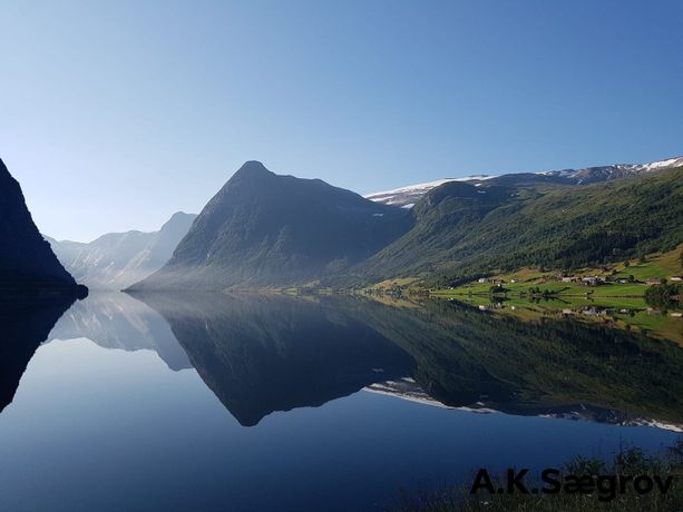 Utsikt over en helt stille fjord som gjenspeiler fjellene, trærne, hus og den blå himmelen 