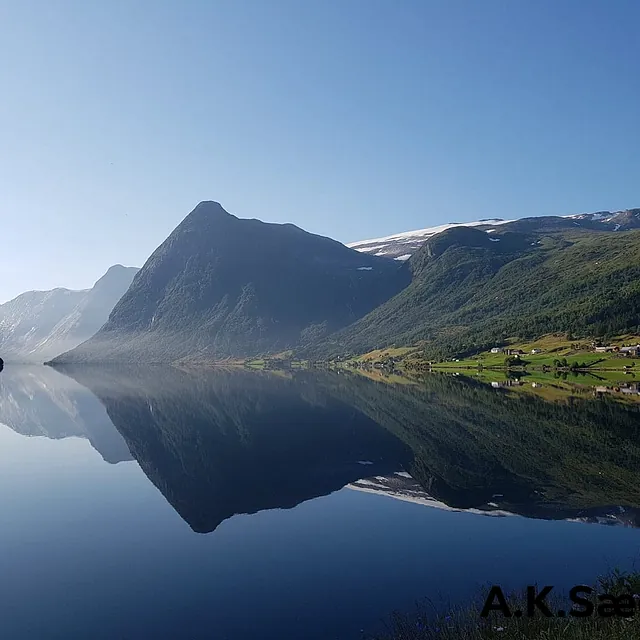 Utsikt over en helt stille fjord som gjenspeiler fjellene, trærne, hus og den blå himmelen 