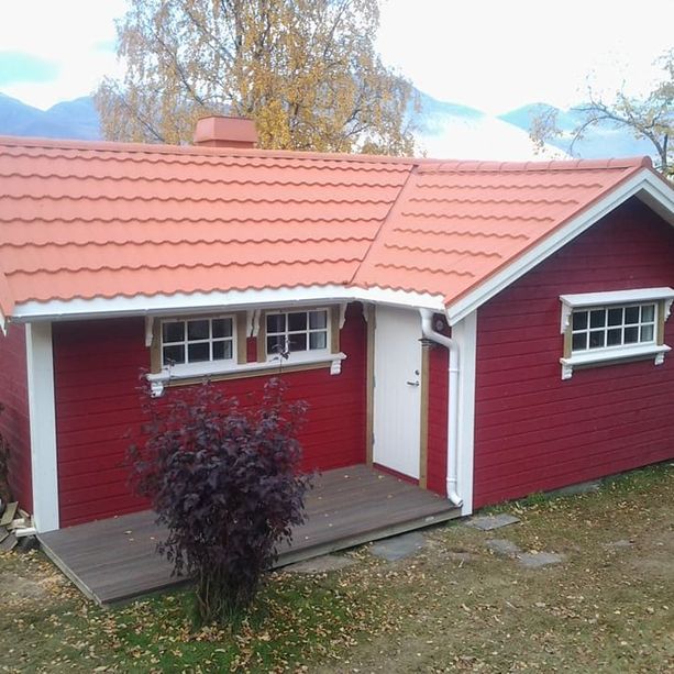 En rød hytte med bølgete takplater og en pipe som stikker opp, hvite, klassiske, detaljerte vinduskarmer og en liten platting som går til inngangsdøren