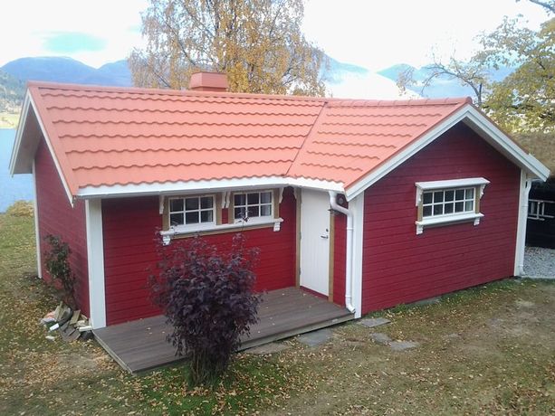 En rød hytte med bølgete takplater og en pipe som stikker opp, hvite, klassiske, detaljerte vinduskarmer og en liten platting som går til inngangsdøren