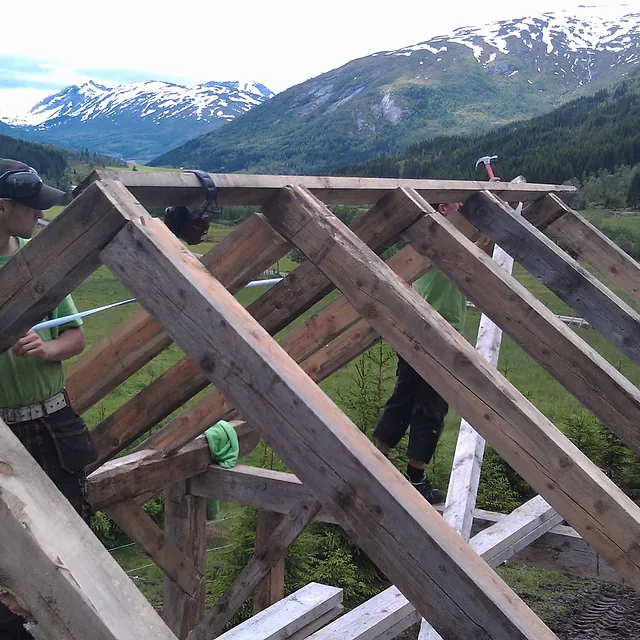 To ansatte som setter opp skjelettet til et hus i et lite bebodd område omringet av fjell 