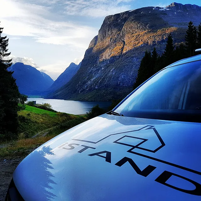 Firmabil som står i forgrunnen av bildet med en fantastisk utsikt bak den av et tre før man ser en fjord og mange fjell med noen skyer på himmelen 