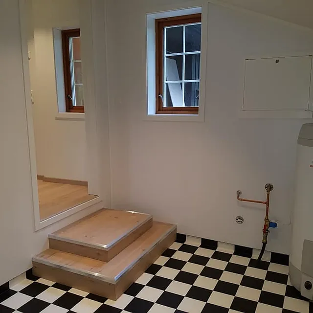 Et vaskerom/bod med en stor vanntank, svart og hvite fliser, vinduer og trapper som leder opp tilbake til resten av huset