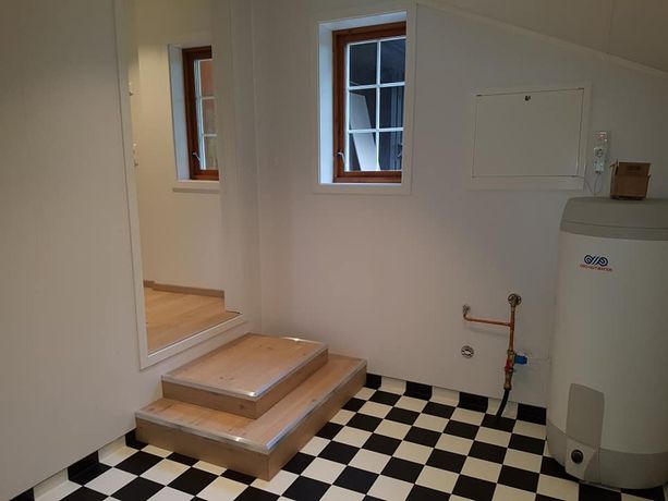 Et vaskerom/bod med en stor vanntank, svart og hvite fliser, vinduer og trapper som leder opp tilbake til resten av huset