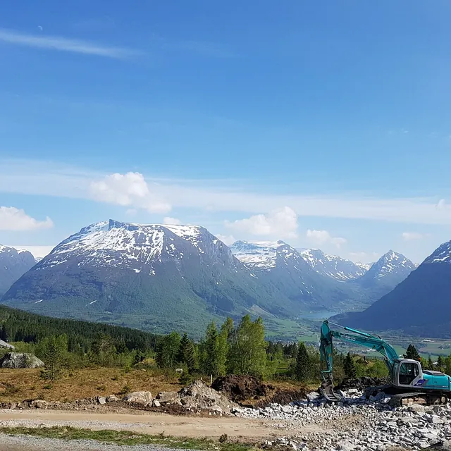 en gravemaskin som graver ut en ny tomt med en utsikt av blå himmel, daler, fjell med snødekte fjelltopper