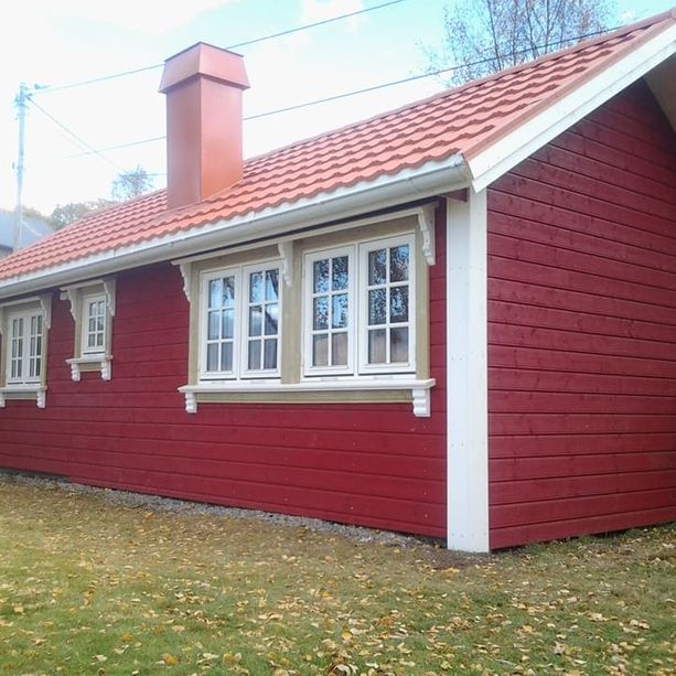 En rød hytte med bølgete takplater og en pipe som stikker opp, hvite detaljerte vinduskarmer, hvite tømmer på hushjørnene samt høstløv på bakken