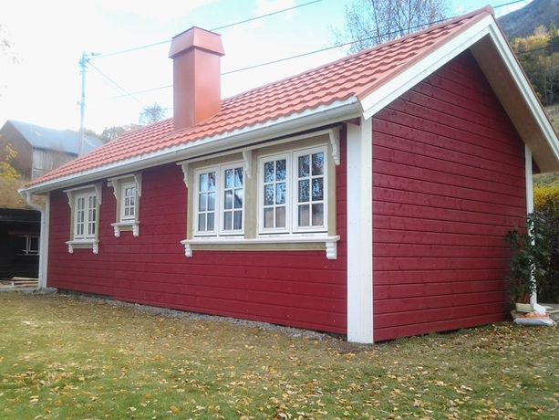 En rød hytte med bølgete takplater og en pipe som stikker opp, hvite detaljerte vinduskarmer, hvite tømmer på hushjørnene samt høstløv på bakken