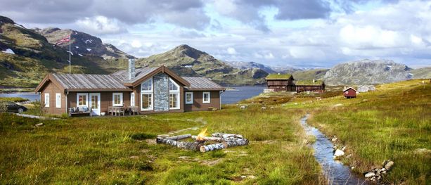 Et hyttefelt ved et vann på fjellet med en bekk som renner forbi en stor hytte med store hvite vindu, bål, parkering rett ved hytten, norske flagg, gamle boder med torvtak og fjell i bakgrunnen