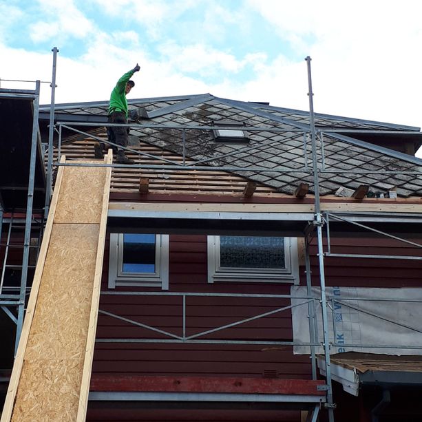 En ansatt som arbeider med å fjerne gamle takplater og sender de ned på en hjemmelagd tre sklie som er lent opp på et stilas på huset