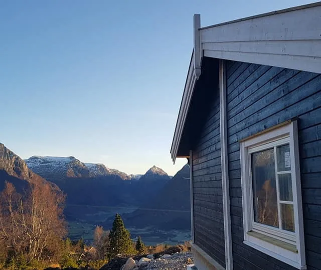 Nærbilde av hjørnet til en svart hytte med hvite vinduskarmer, takskjegg og en fantastisk utsikt ut over en dal og fjelltopper i bakgrunnen