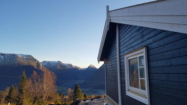 Nærbilde av hjørnet til en svart hytte med hvite vinduskarmer, takskjegg og en fantastisk utsikt ut over en dal og fjelltopper i bakgrunnen