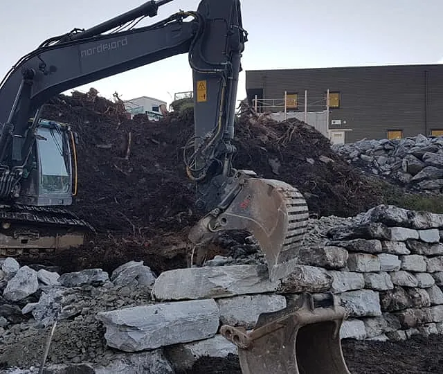 En gravemaskin som legger ned store steiner i en høy grøft hvor man ser et hus i bakgrunnen