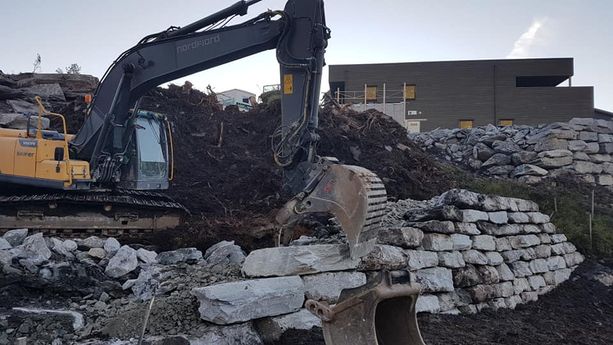 En gravemaskin som legger ned store steiner i en høy grøft hvor man ser et hus i bakgrunnen