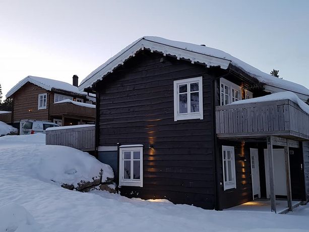 Mørk hytte med detaljert dekortert hvitt takskjegg, hvite vinduskarmer, en balkong og hvit inngangsdør i et vinterlandskap dekket i snø i et hyttefelt