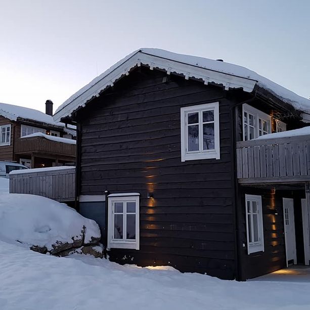 Mørk hytte med detaljert dekortert hvitt takskjegg, hvite vinduskarmer, en balkong og hvit inngangsdør i et vinterlandskap dekket i snø i et hyttefelt
